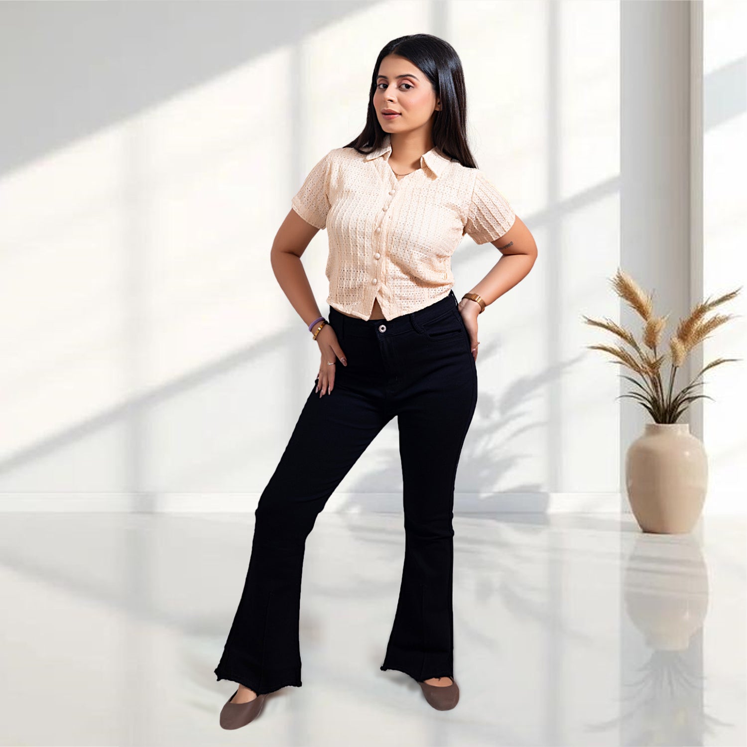 Woman wearing a light-colored blouse and black pants standing in a bright room with a plant in the background.